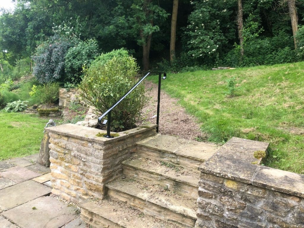 Stone steps with a metal railing lead up to a grassy area surrounded by dense greenery and trees. A small stone wall borders the steps, with shrubs and foliage beyond.