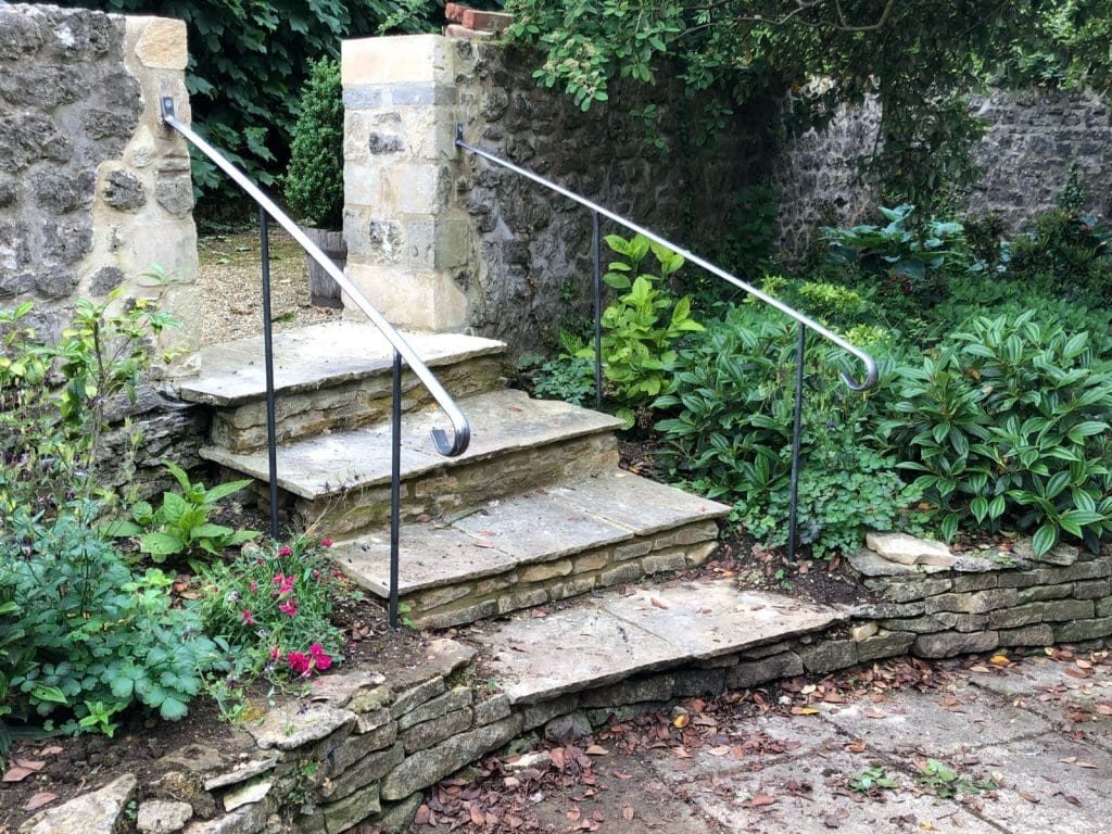 Stone steps with metal railings lead to a garden area, surrounded by stone walls and lush greenery. Pink flowers and various plants are visible, creating a serene and inviting outdoor space.