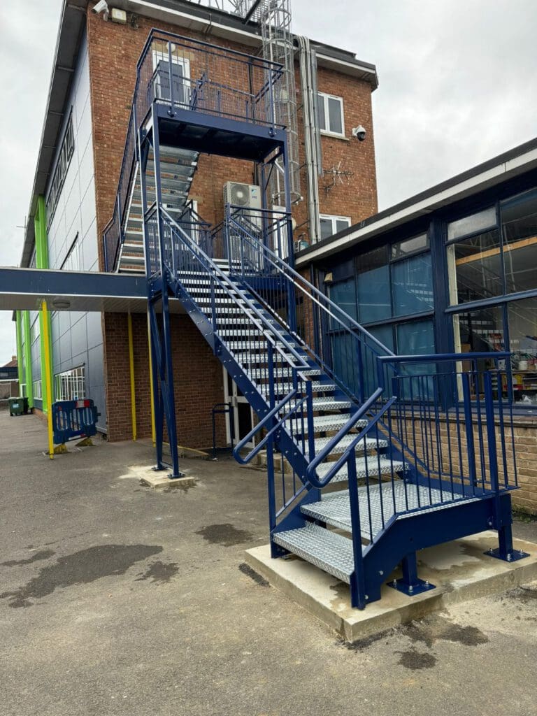 A blue metal staircase outside a brick and metal building. The stairs lead to a second-story door. The surrounding area is paved, and there are windows on the building. The sky is overcast.