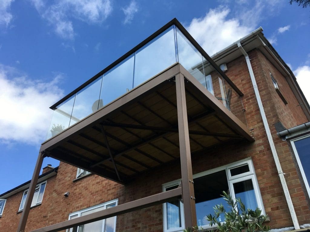 A view from below of a brick house with a glass-railed balcony extending outward. The balcony is supported by metal beams. The sky is clear with a few clouds.