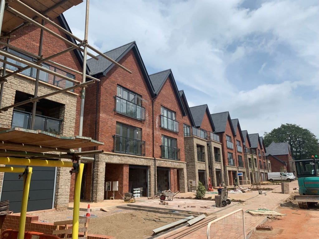 A row of newly constructed red-brick townhouses with large windows and gabled roofs under a partly cloudy sky. Construction materials and equipment are visible on the ground, indicating ongoing development work.