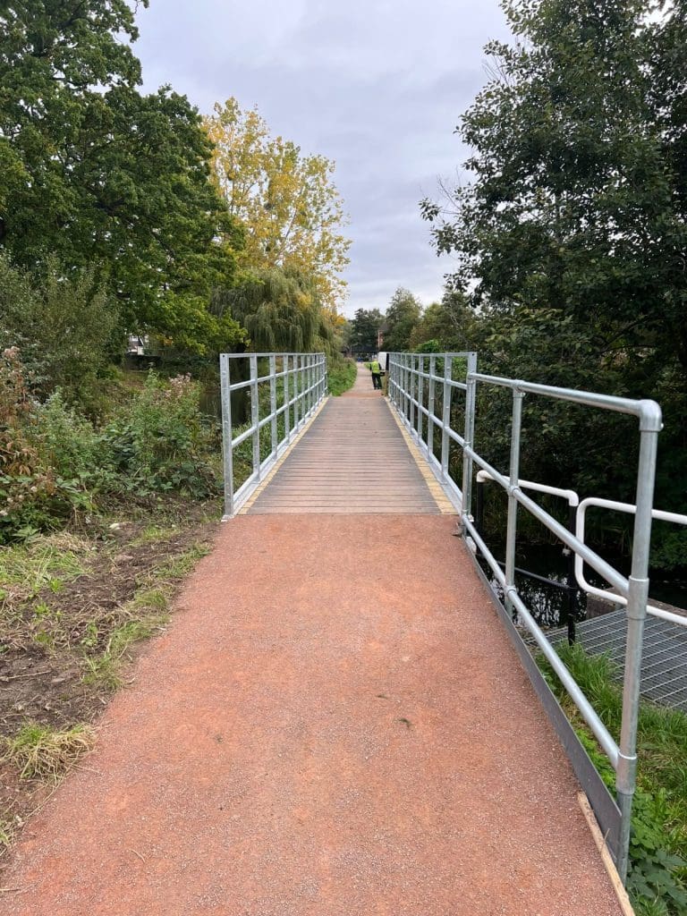 A narrow metal and wood footbridge extends across a small waterway, flanked by green foliage. The path leading to the bridge is reddish-brown, and the sky is overcast, with trees visible in the background. A person is walking in the distance.