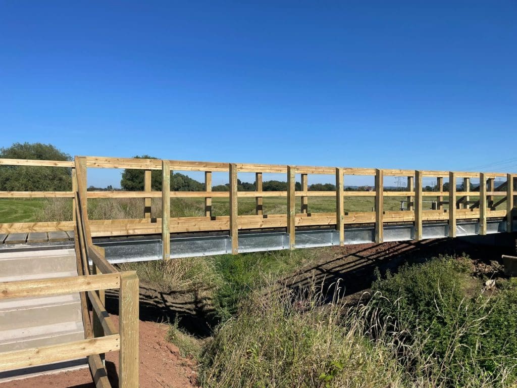 A wooden pedestrian bridge with metal supports spans over grassy terrain. Clear blue skies and green fields are visible in the background.