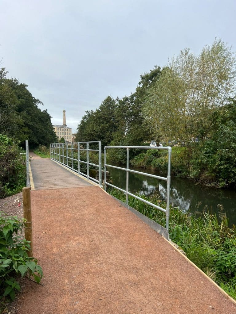 A narrow path with a metal railing runs alongside a calm river, surrounded by lush greenery. A large brick building with a tall chimney is visible in the distance under an overcast sky.