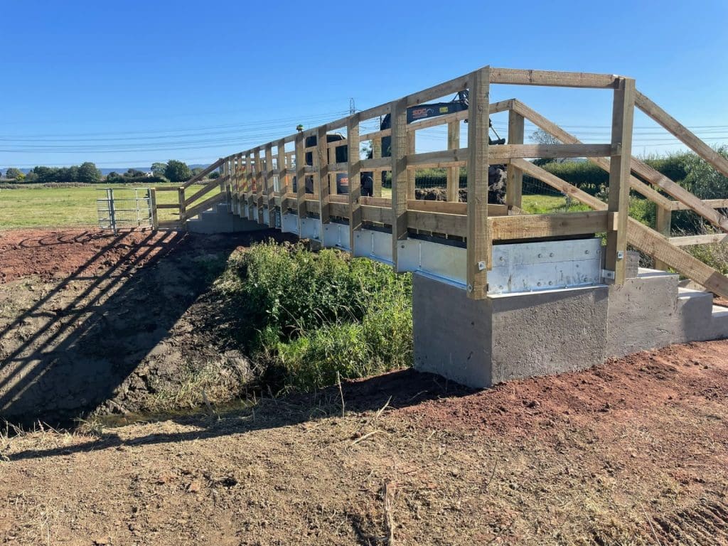 A wooden footbridge with handrails spans over a small ditch in a rural area. The sky is clear and blue, and the surrounding landscape includes green fields and distant trees. The ground has patches of red earth and vegetation near the bridge.