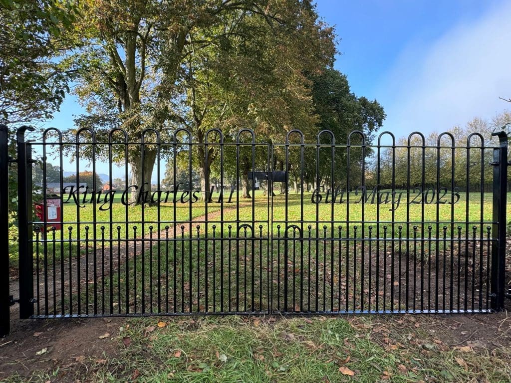 A black metal gate with the inscription "King Charles III Coronation 6th May 2023" stands open in a park. Trees with green leaves surround the area, and a clear blue sky is visible above.