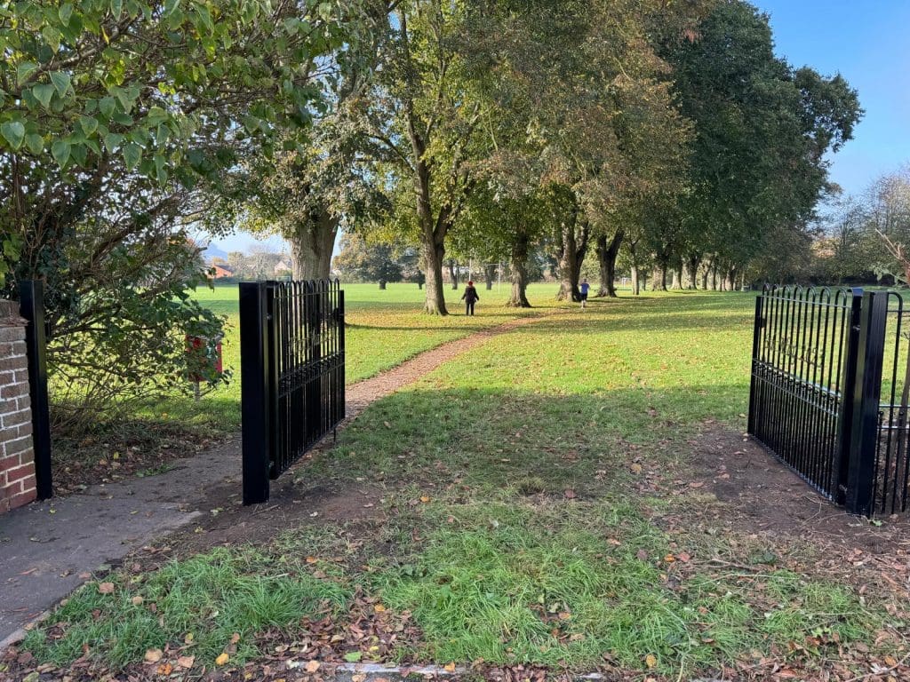 Open black gates lead to a grassy park area with a path lined by tall trees. A person walks on the path in the distance under a clear blue sky.