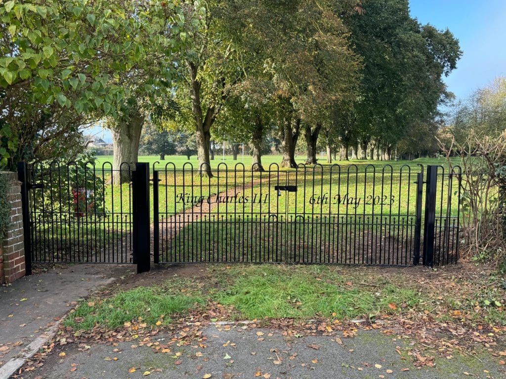 A black metal gate opens to a lush park with large trees and fallen leaves. A path leads through the green grass. Inscribed on the gate are the words "Rise Gardens 11th - 13th May 2023." Brick wall and sidewalk are visible.