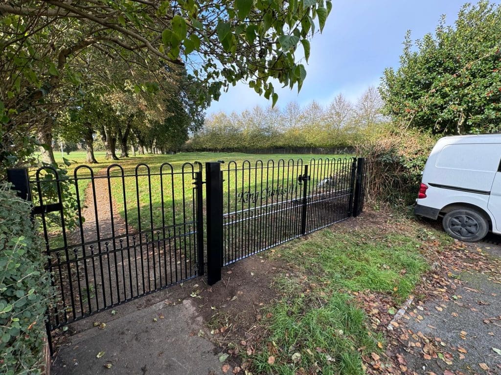 A black metal gate leads to a grassy field surrounded by trees. The gate is partway open, with a small white van parked on the right side of a paved road. The sky is clear and sunny, and there are bushes lining the area.