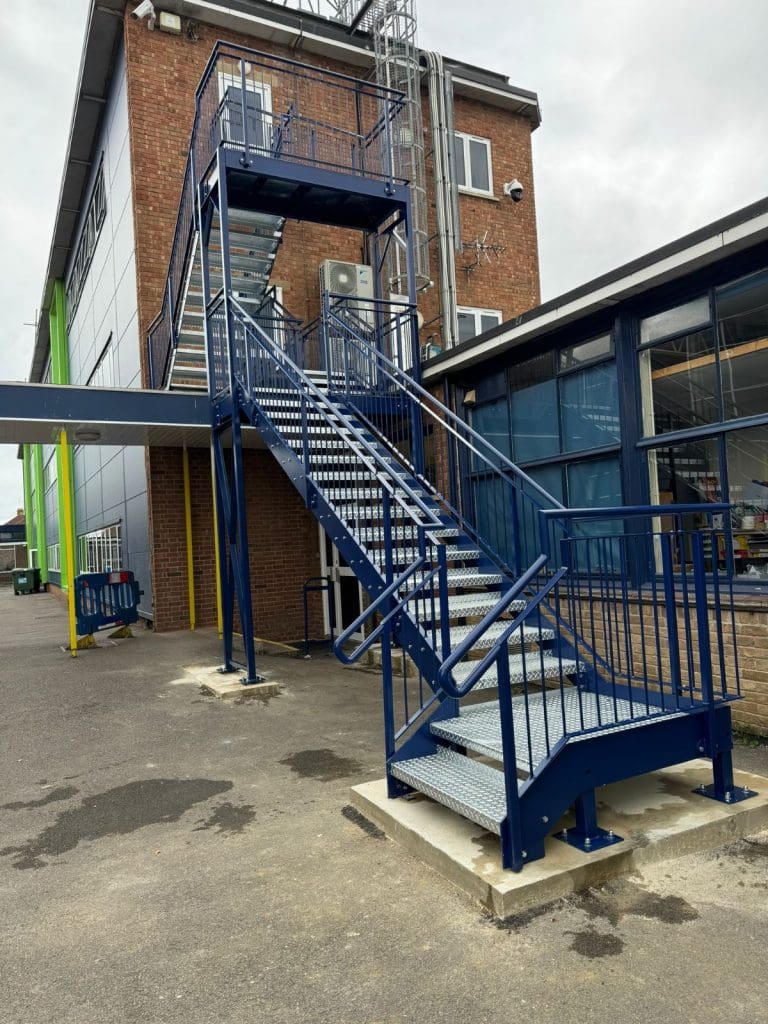 A blue metal staircase leads up the side of a red brick building with two levels. The structure is supported by metal beams, next to a gray wall extension and an enclosed glass area. The surrounding ground is paved.