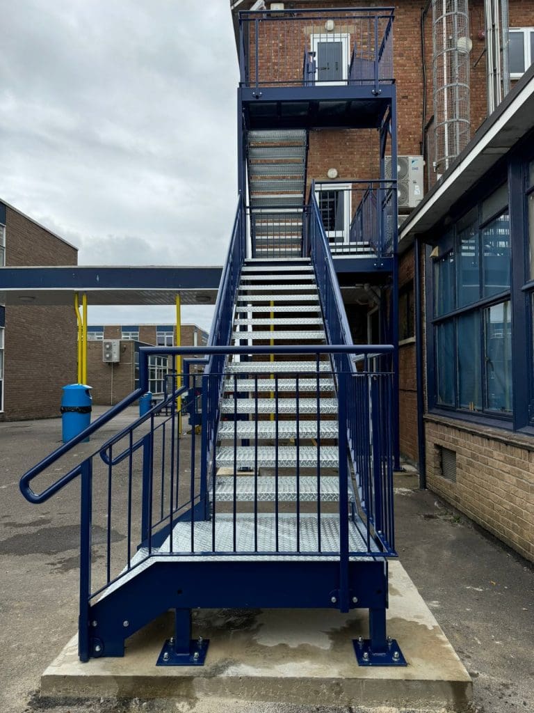 A blue metal outdoor staircase with safety railings, leading up to a balcony attached to a brick building. The sky is overcast, and there are adjacent buildings and a blue trash bin visible in the background.