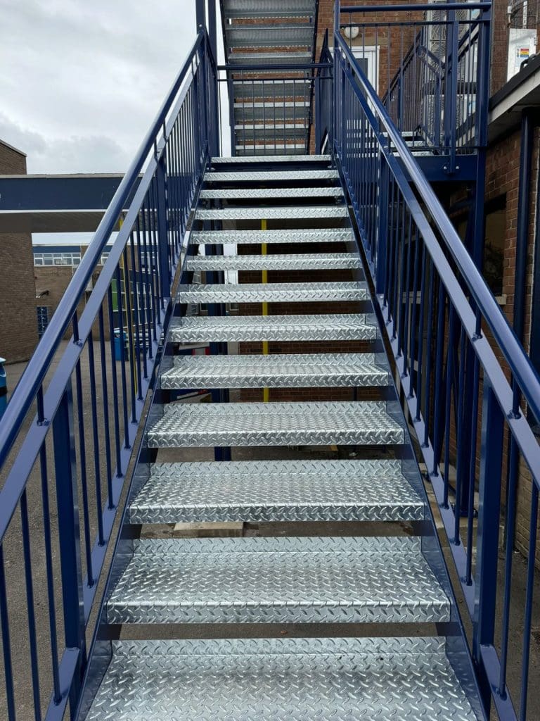 A metal staircase with blue railings leads upward against a brick building. The steps are made of diamond plate metal. The sky is overcast, and nearby structures are visible in the background.