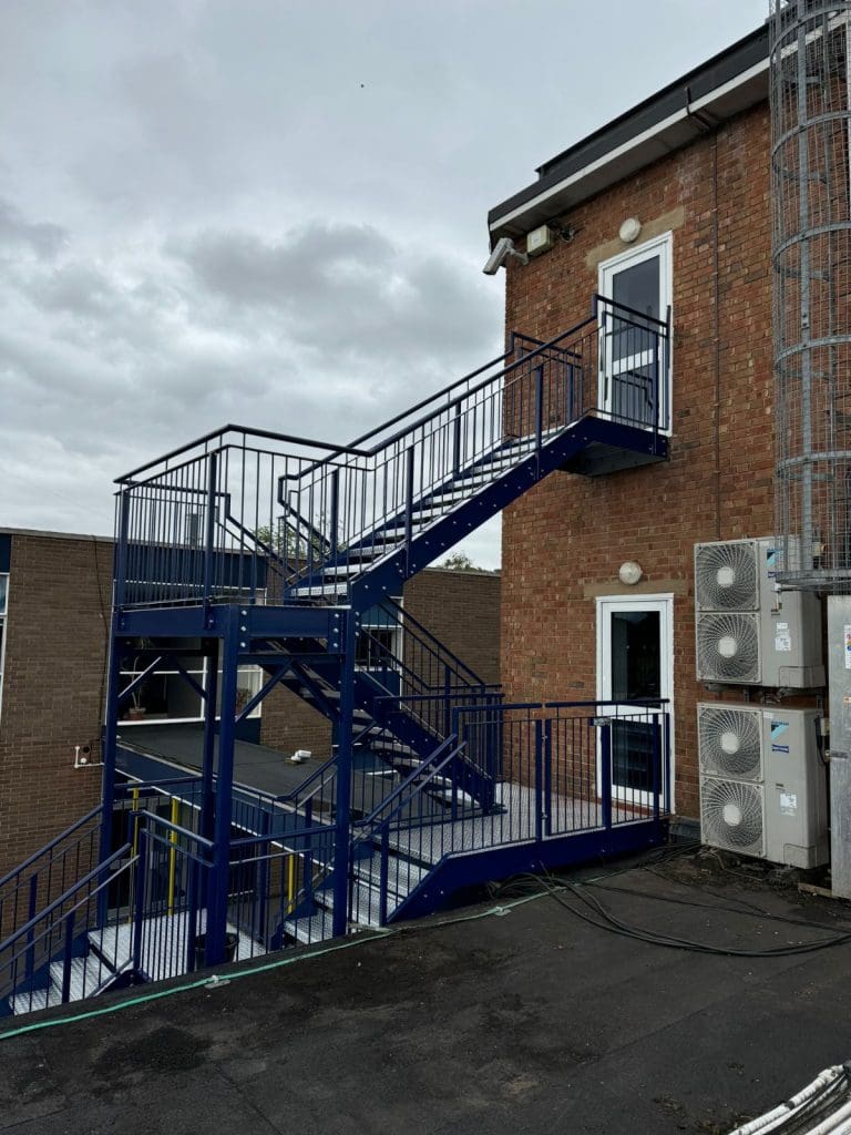 A metal outdoor staircase with blue railings leads to a brick building. The sky is cloudy, and several air conditioning units are mounted on the wall beside the staircase.