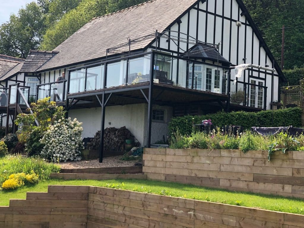 A modern house with a black and white exterior features large windows and a balcony. It's built on a sloped green lawn with stacked wooden terraces and surrounded by lush greenery and flowering plants.