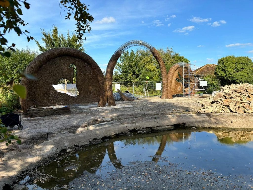 A scenic outdoor installation featuring large, woven straw arches. The arches stand on a grassy area by a shallow pond under a clear blue sky. Scaffolding and a pile of rocks are nearby, with trees and bushes in the background.