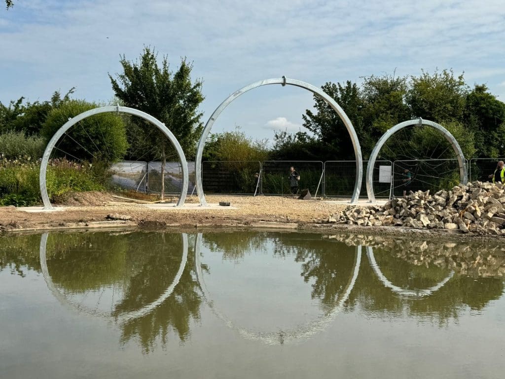 Three large metal arches stand beside a reflective pond, with trees in the background and construction materials nearby. A person walks past a fenced area under a cloudy sky.