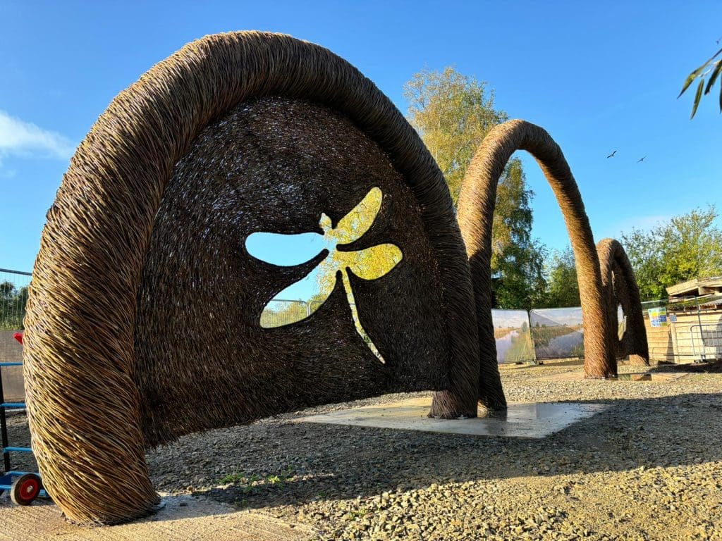 A large, woven sculpture with arching forms and a dragonfly cutout stands against a clear blue sky. It casts a shadow on the ground, with trees and birds visible in the background.