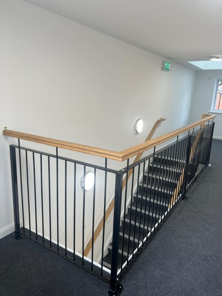 A modern, indoor staircase with black metal railings and wooden handrails. The staircase is carpeted in dark gray and illuminated by circular wall lights. An emergency exit sign is visible above a door at the end of the hallway.