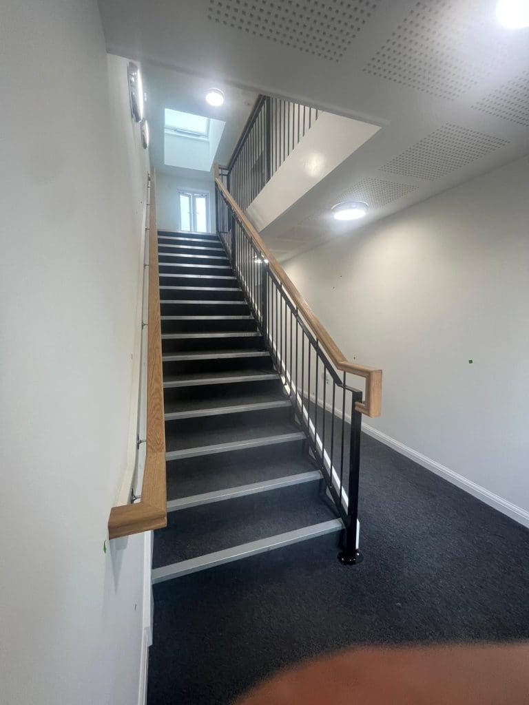 A staircase with wooden handrails, black metal railings, and gray carpeted steps leading to a bright window at the top. The walls and ceiling are white, with circular ceiling lights illuminating the space.