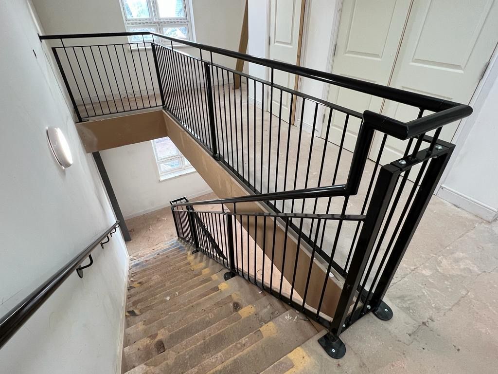 A stairwell with black metal railings and a concrete floor. The stairs have a strip of yellow tape on the edge of each step. Walls are light-colored, with a round light fixture and a window on the left side.