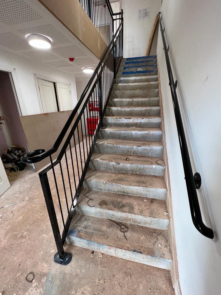 Staircase under construction with a metal railing on the left side and a partially covered blue tarp on some steps. The area appears unfinished, with construction materials scattered around.