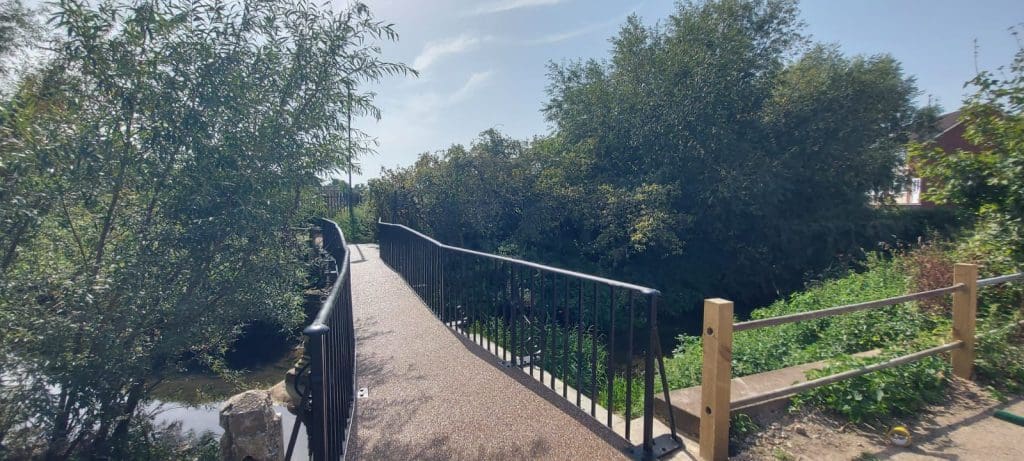 A narrow footbridge with black railings spans a small stream, surrounded by lush green trees and foliage. The sky is clear and blue, indicating a sunny day. A wooden post with horizontal beams stands on the right.