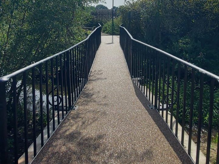 A narrow footbridge with metal railings crosses over a small stream. The bridge is surrounded by lush green trees and shrubs under a bright blue sky.