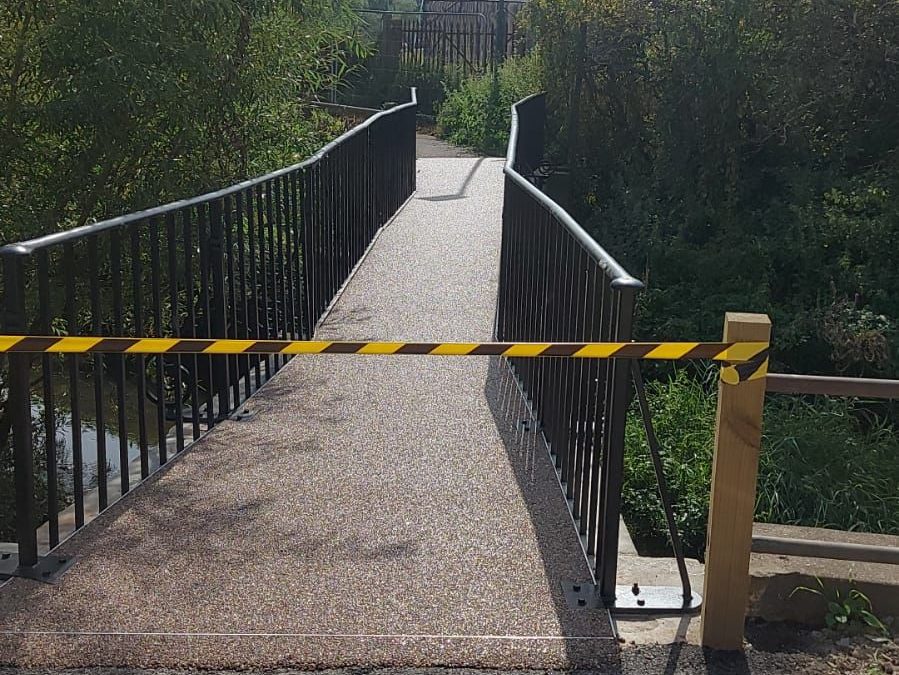 A pedestrian bridge is blocked with black and yellow caution tape. The bridge has metal railings and crosses over a small stream surrounded by greenery. A narrow path extends beyond the bridge.