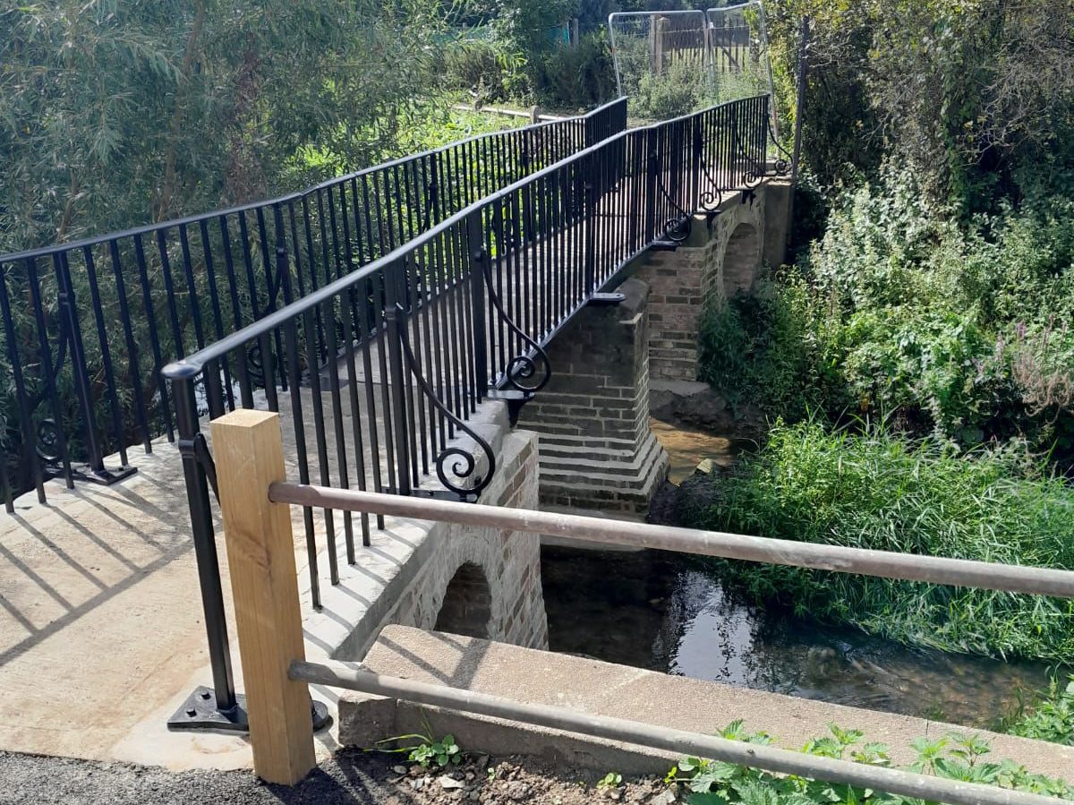 A small pedestrian bridge with black metal railings spans over a narrow stream. The bridge is supported by brick pillars and surrounded by green foliage and tall grasses on either side.