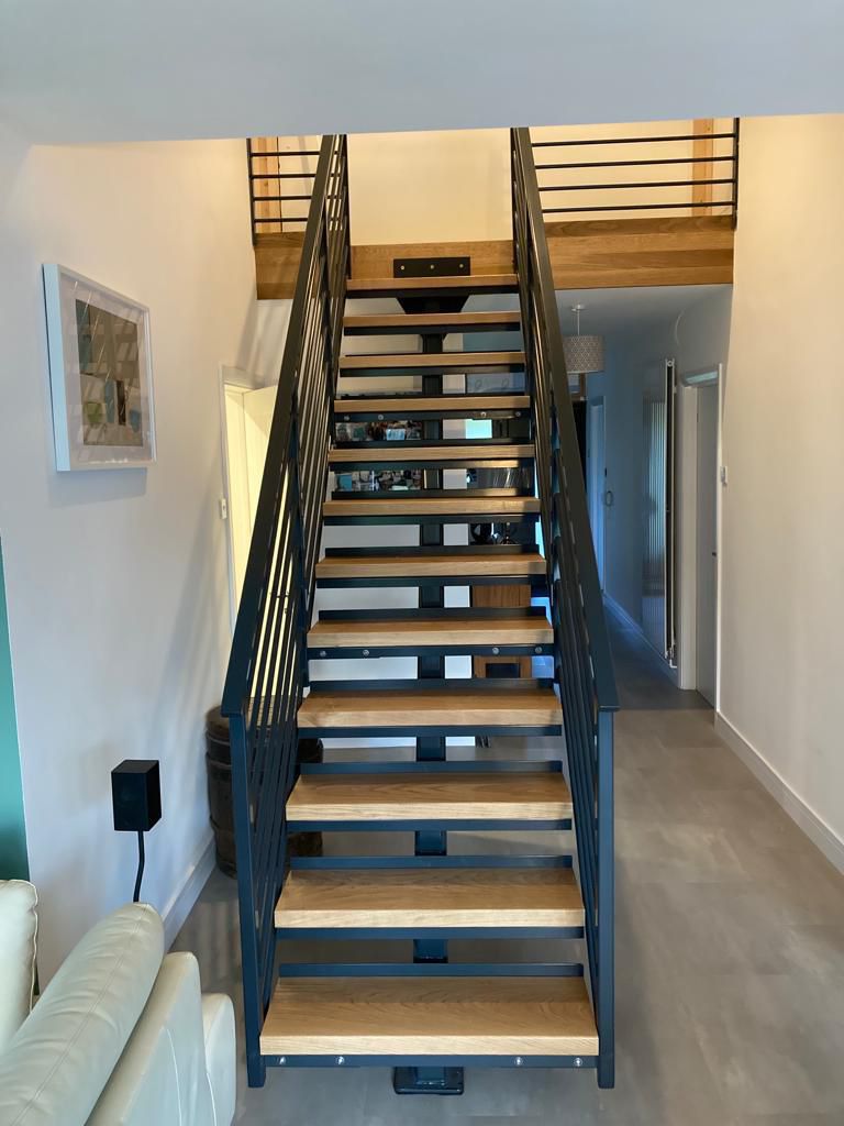 A modern, straight staircase with wooden steps and black metal railings in a home's interior. The staircase connects two levels, with artwork on the wall and a hallway visible on the right side.