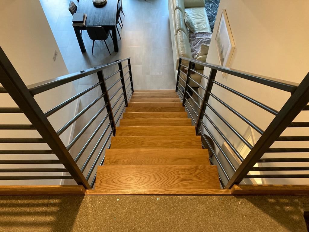 A straight wooden staircase with metal railings on both sides leads down to a modern living area. The living space features a dining table, chairs, and a couch, all set on a wood-paneled floor.