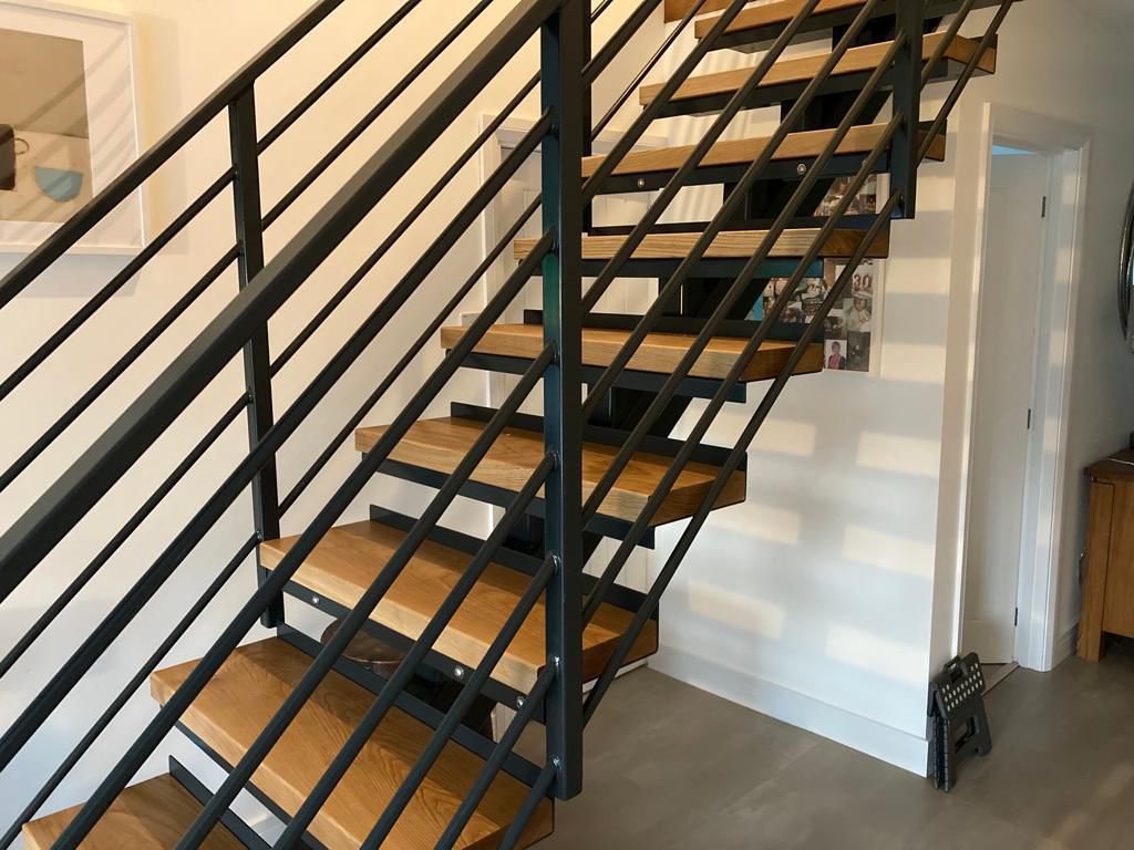 A modern staircase with wooden steps and black metal railings in a well-lit interior. The walls are white, and there's framed artwork near the stairs. A small, dark object rests on the floor beneath the staircase.