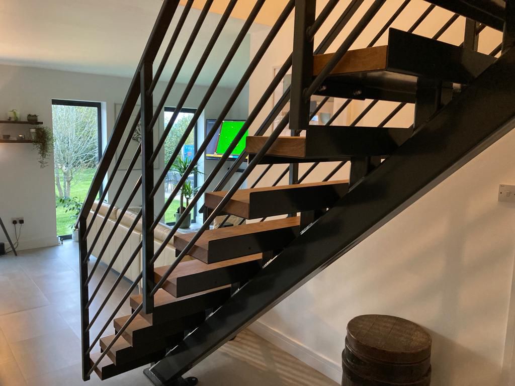 A modern interior features a wooden staircase with metal railings. The room has tile flooring, large windows with a view of greenery outside, and shelves with plants on the wall. A round wooden table is placed near the staircase.